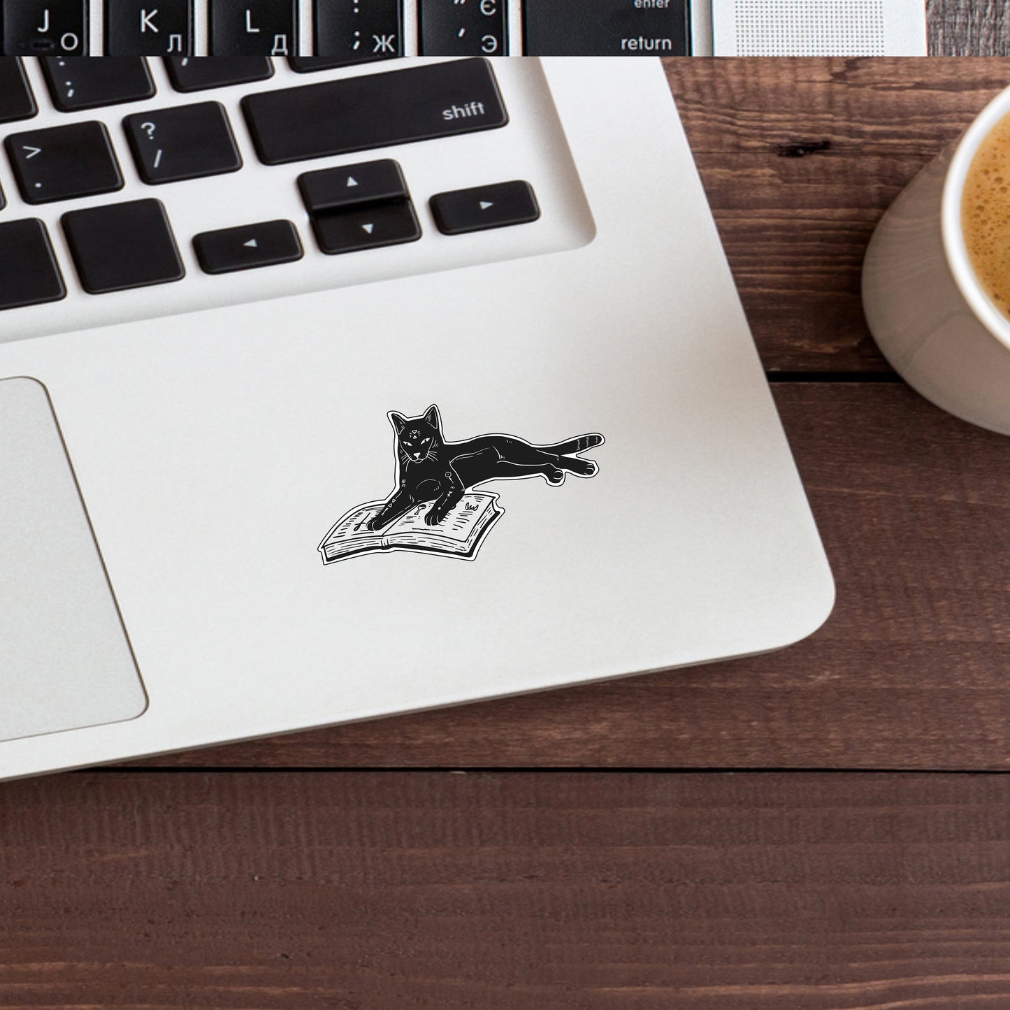 a cat sticker on a laptop next to a cup of coffee